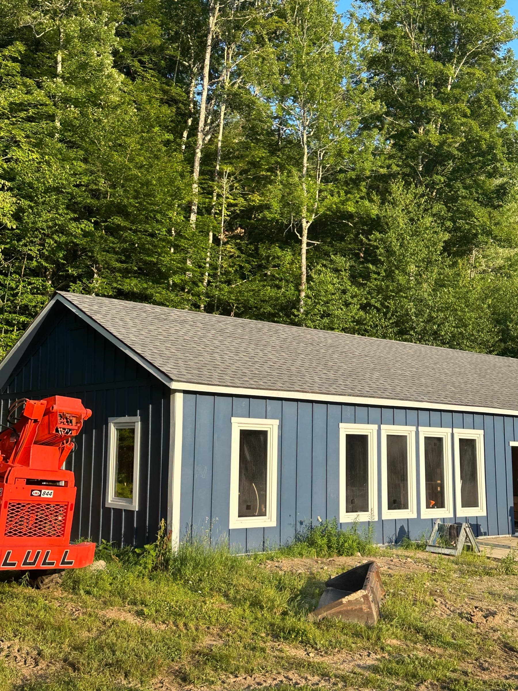Single-story build clad in slate blue board-and-batten siding with cream trim, against a Vermont treeline